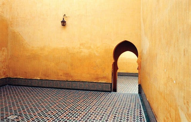 A Moroccan courtyard with an arched doorway and yellow plaster walls, featuring a patterned tile floor and a single wall-mounted lamp.