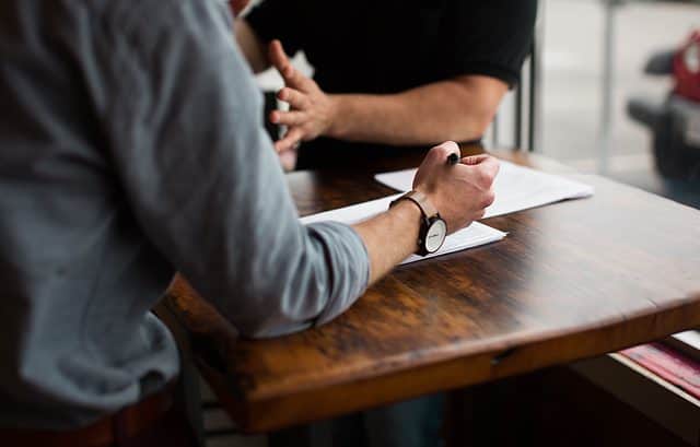 Two people sit at a wooden table, one gesturing while the other holds a pen with papers spread out in front of them, discussing BEPS and CbCR guidelines.