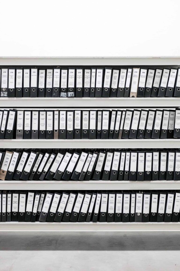 A neatly organized shelf holds numerous black ring binders, including the master file, all labeled and arranged in rows.