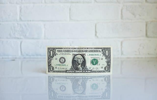 A single U.S. one-dollar bill stands upright on a reflective white surface against a white brick wall background, embodying the essence of economics in the news amid discussions of groundbreaking materials like graphene.