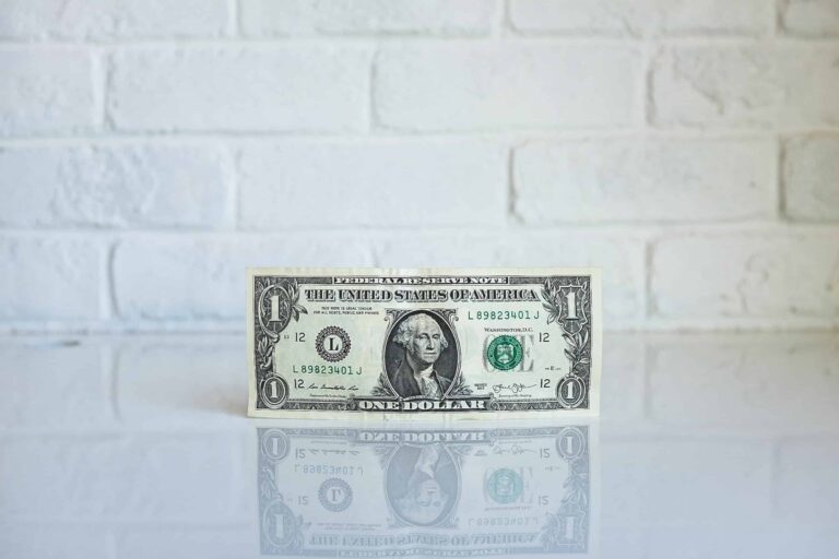 A single U.S. one-dollar bill stands upright on a reflective white surface against a white brick wall background, embodying the essence of economics in the news amid discussions of groundbreaking materials like graphene.