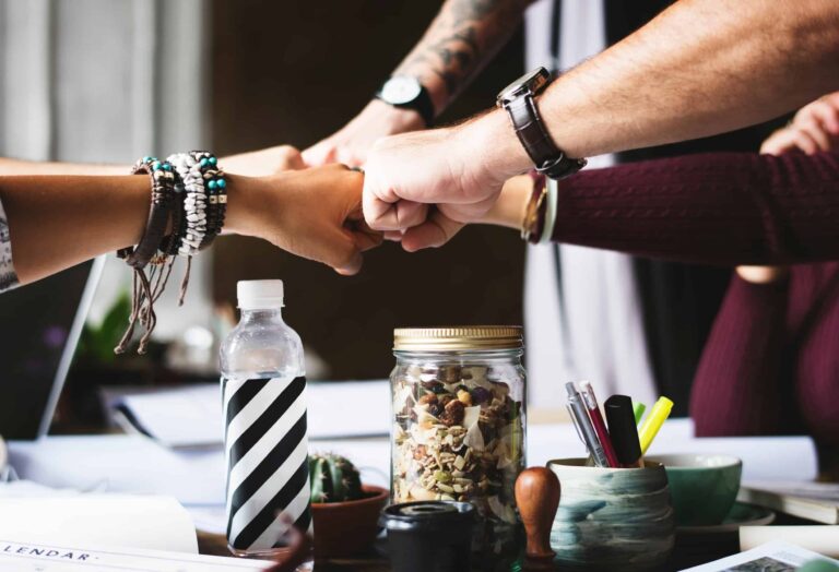 Four people perform a fist bump over a jar on a cluttered desk, showcasing teamwork in their casual office setting. The camaraderie is palpable as they tackle their roles, possibly even aspiring to the Transfer Pricing Consultant position within the vibrant workplace.