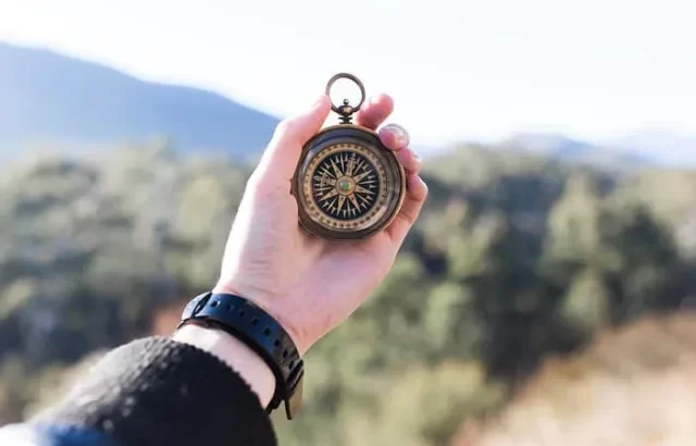 A person holds a brass compass outdoors with a blurred landscape of trees and mountains in the background, reminiscent of navigating through the complexities of a benchmarking study on transfer pricing.