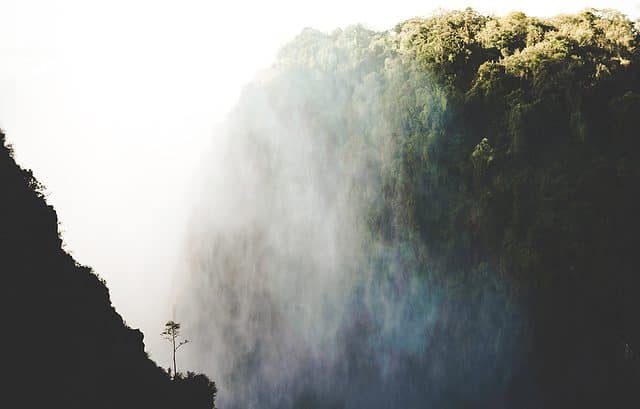 Misty waterfall cascading down a steep, forested cliffside with sunlight illuminating the mist, creating a bright contrast against the greenery, much like navigating Zambia's new transfer pricing regulations amidst evolving economic landscapes.