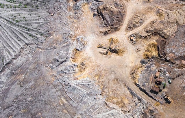 Aerial view of a large mining site with exposed earth, machinery, and tracks. The landscape includes different shades of brown and gray, with some vegetation in the top left corner, hinting at the potential for intra-group funding to further develop the area.