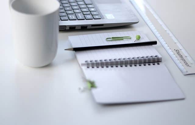 A laptop, a white mug, a ruler, an open spiral notebook with a pencil and paperclip, and a pen on a white desk suggest meticulous attention to reportable arrangements.