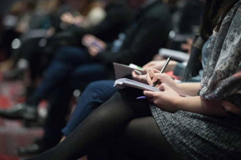 People seated in rows, taking notes with notebooks and pens during an event where speakers discuss new taxing rights.