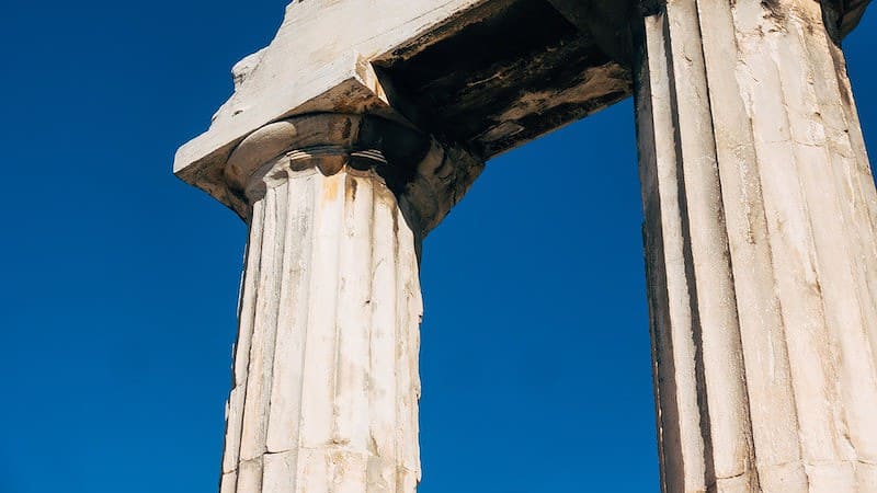 Two ancient stone columns, known as Pillar One and Pillar Two, with weathered surfaces stand under a clear blue sky.