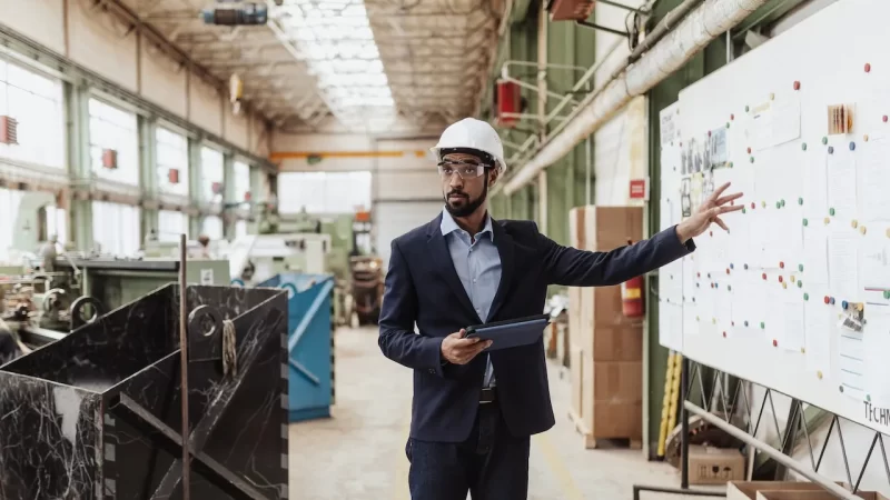 Man in safety gear gestures toward a whiteboard in an industrial facility, holding a tablet. He is surrounded by machinery and large open workspace, illustrating operational exposure.