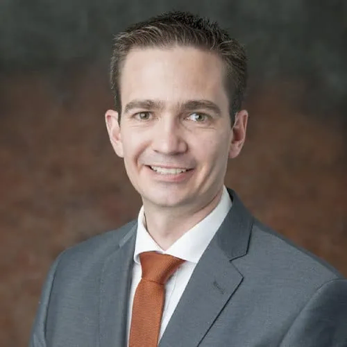 A man in a gray suit, white shirt, and orange tie smiles at the camera against a brown and gray background, reflecting the professionalism expected in transfer pricing roles.