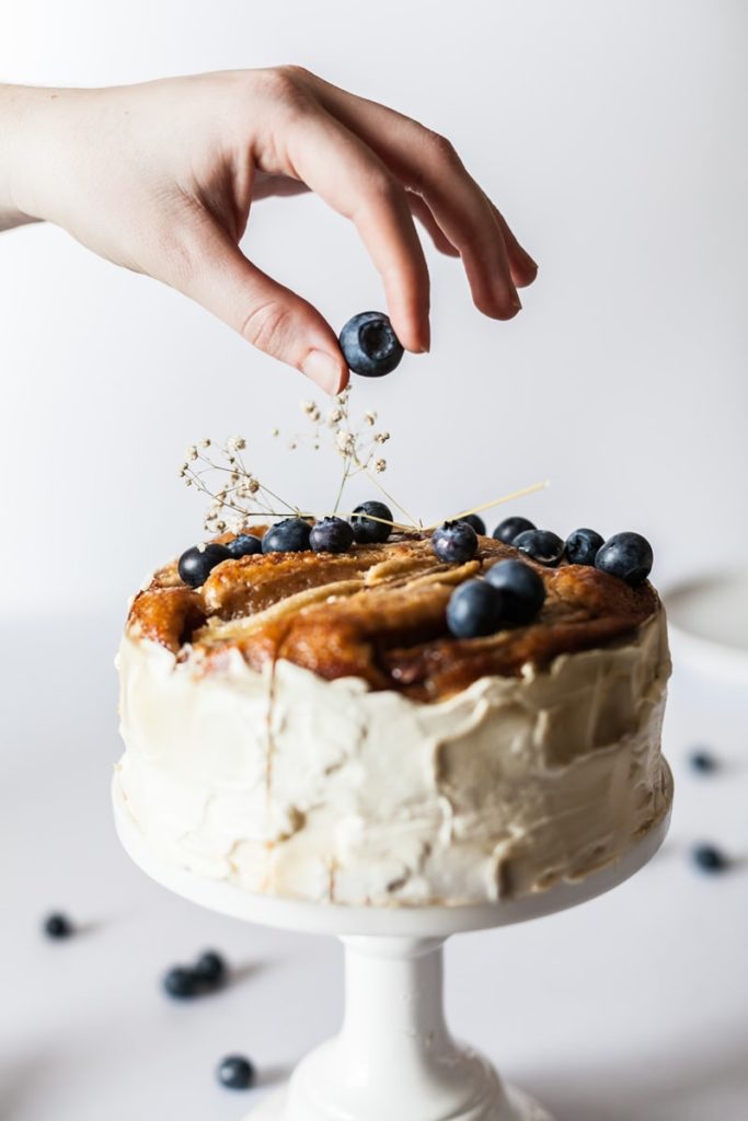 A hand places a blueberry on top of a frosted cake decorated with blueberries and small flowers, set on a white cake stand—an arrangement as precise as transfer pricing in global finance.