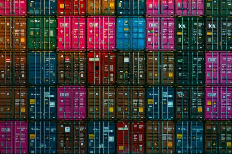 A large stack of colorful shipping containers is arranged in a tightly packed grid pattern, illustrating the complexity of global supply chains through visible doors and labels on each container.