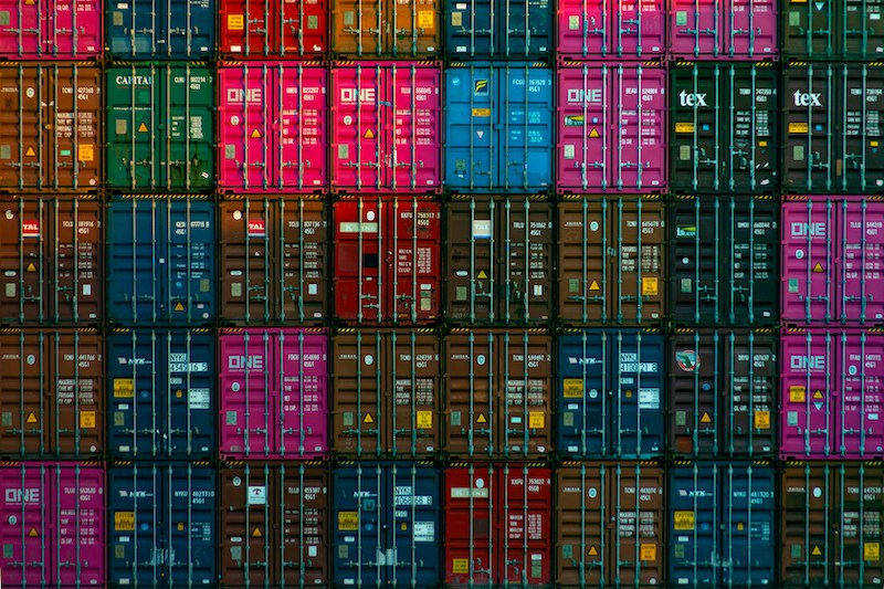 A large stack of colorful shipping containers is arranged in a tightly packed grid pattern, illustrating the complexity of global supply chains through visible doors and labels on each container.